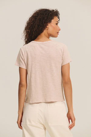 A woman with curly brown hair, shown from the back against a plain background, wears the MARIKA TEE by Velvet by Graham & Spencer in light pink cotton slub boxy fit with cream pants.