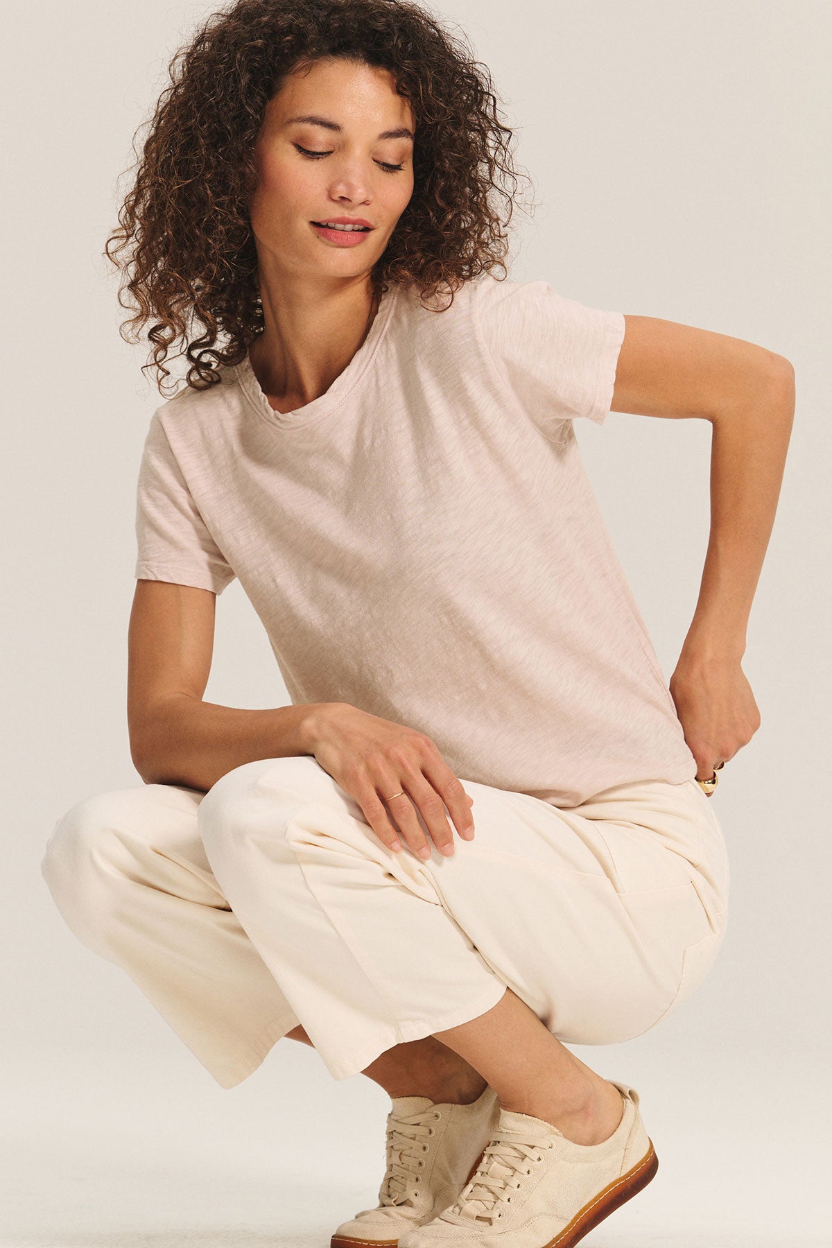 A woman with curly hair wears a light pink MARIKA TEE by Velvet by Graham & Spencer in cotton slub with a boxy fit, paired with white pants and beige sneakers, while squatting against a plain background.-40417104953537