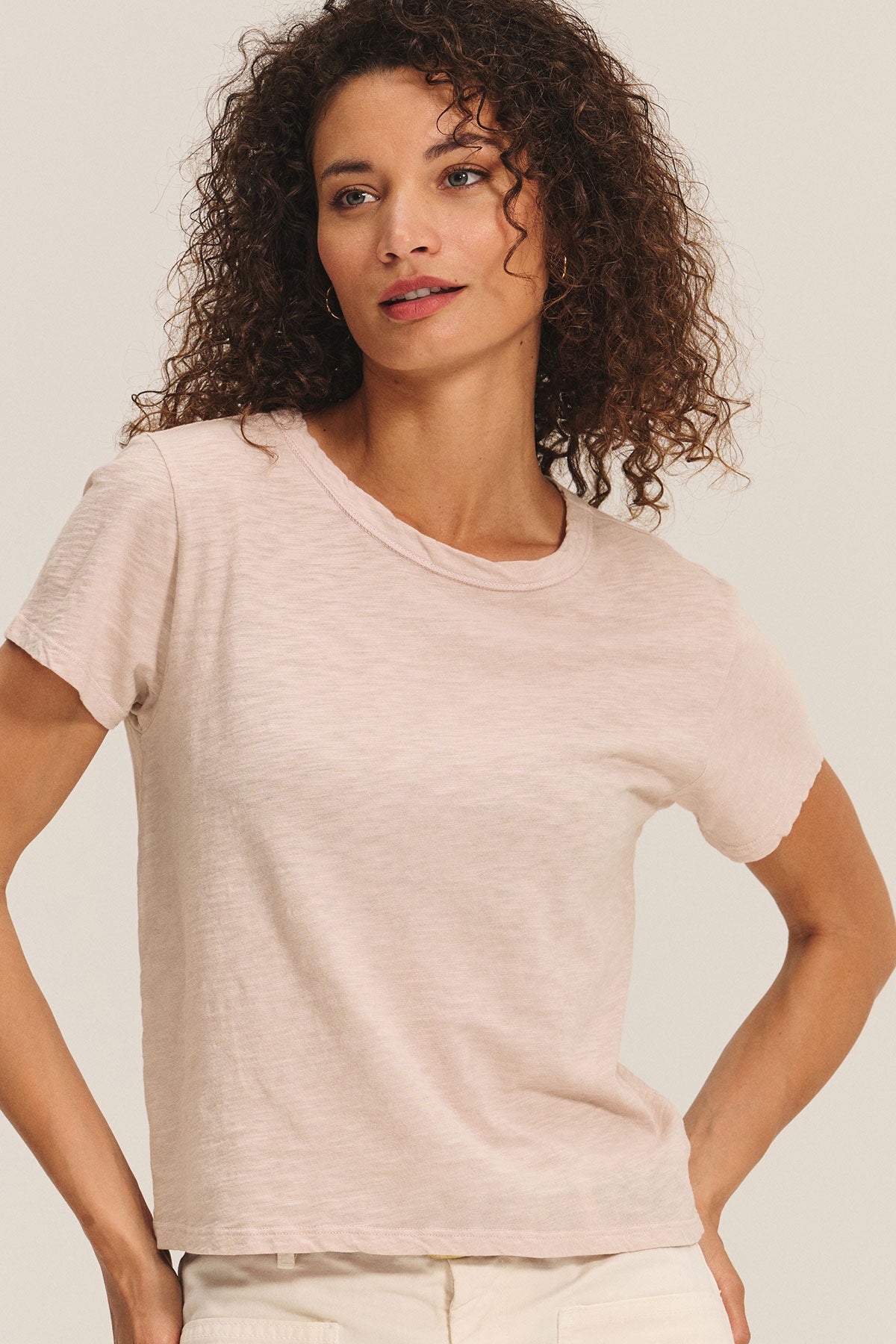 A woman with curly hair wears the light pink MARIKA TEE by Velvet by Graham & Spencer, featuring a cotton slub fabric and boxy fit, paired with cream pants against a plain background.-40417104888001