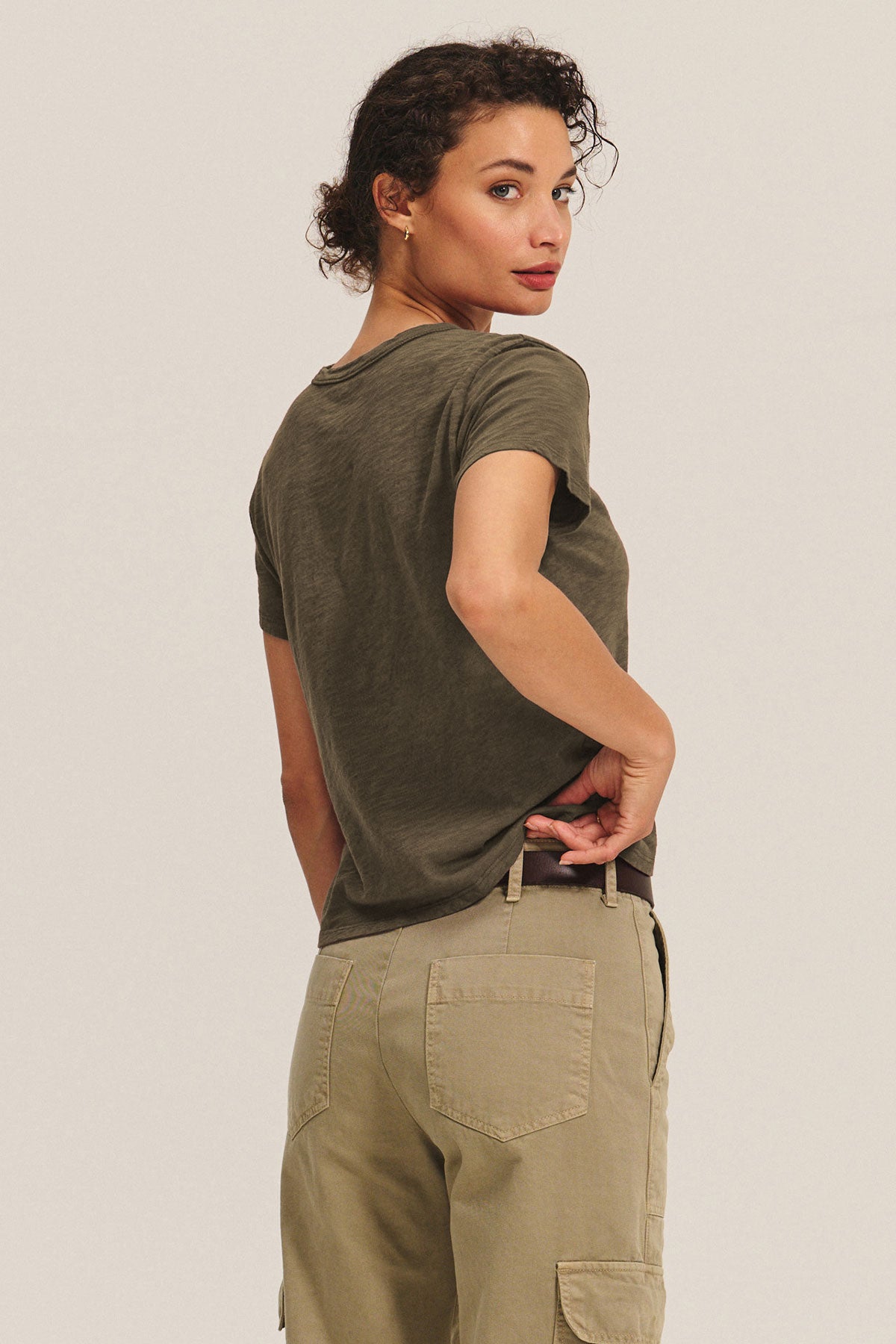 Wearing a cotton slub MARIKA TEE by Velvet by Graham & Spencer and tan cargo pants, a woman stands facing backward, glancing over her shoulder with one hand tucked into her waistband.