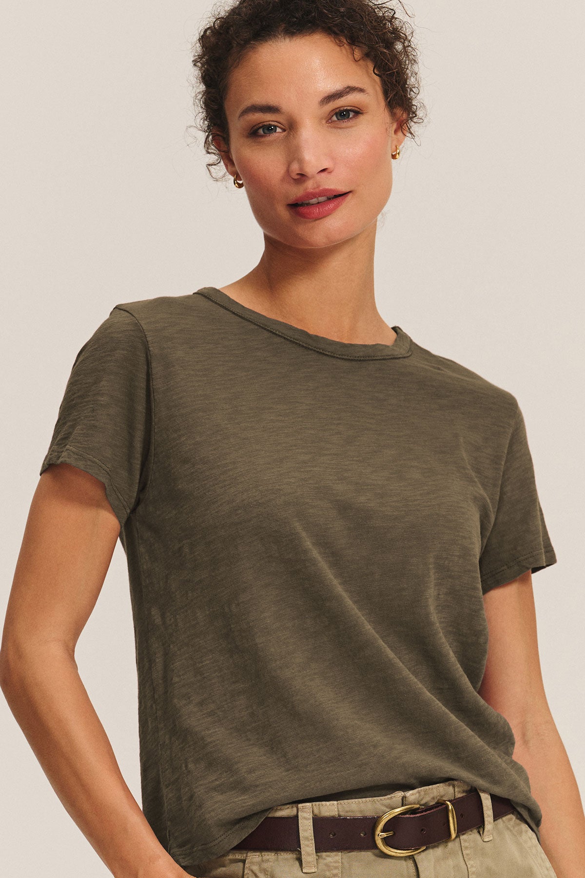 A woman wearing the Velvet by Graham & Spencer MARIKA TEE, a boxy fit olive green cotton slub t-shirt, paired with khaki pants and a brown belt, stands against a plain light background.-40417104527553