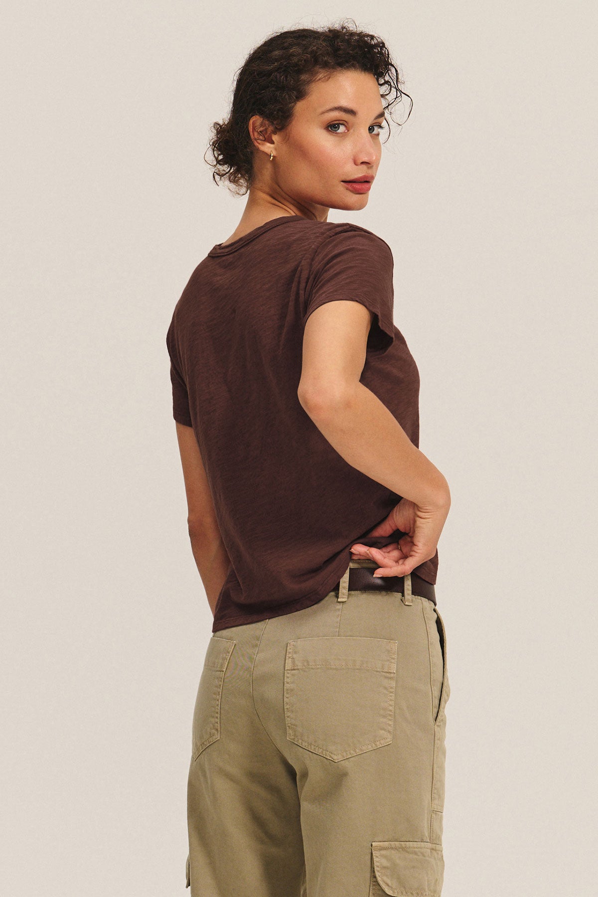 A woman stands with her back turned, looking over her shoulder, wearing a boxy fit MARIKA TEE by Velvet by Graham & Spencer in brown cotton slub and khaki cargo pants.-40417104756929