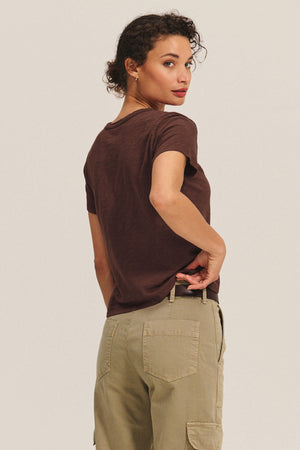 A woman stands with her back turned, looking over her shoulder, wearing a boxy fit MARIKA TEE by Velvet by Graham & Spencer in brown cotton slub and khaki cargo pants.