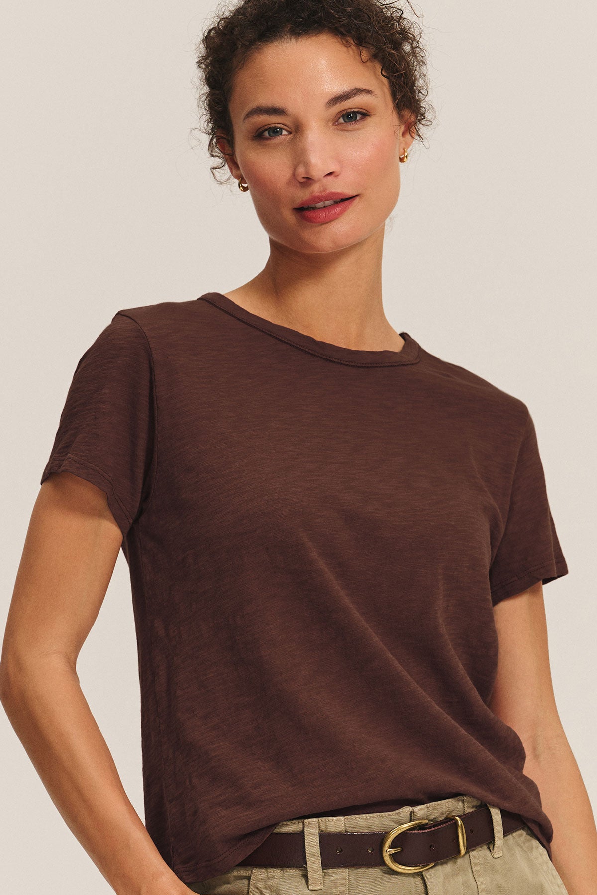 A woman in a short-sleeved MARIKA TEE by Velvet by Graham & Spencer, featuring a boxy cotton slub fit, pairs it with beige pants and a brown belt while standing against a neutral background.