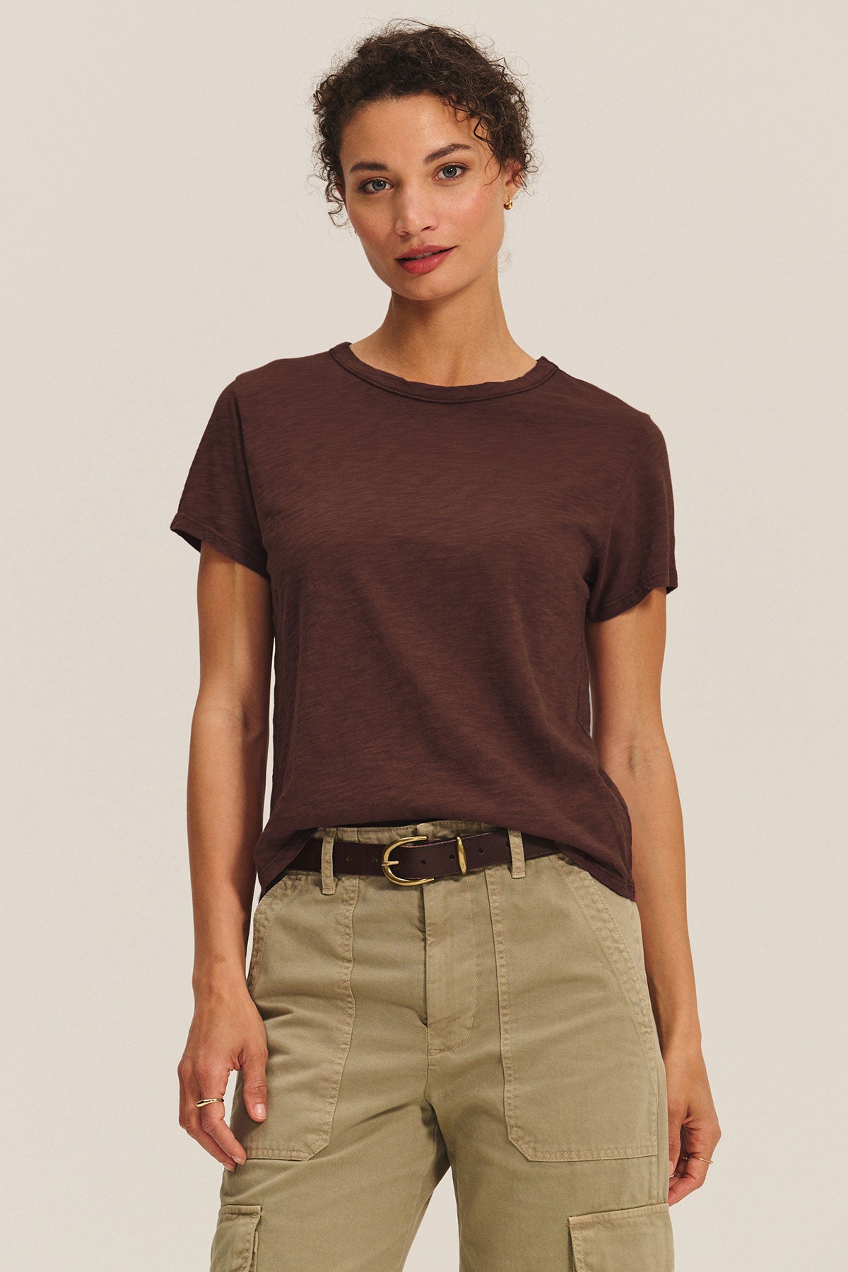 A woman stands against a plain background wearing the Velvet by Graham & Spencer MARIKA TEE in brown, styled in a boxy fit and tucked into khaki cargo pants with a black belt.-40417104691393