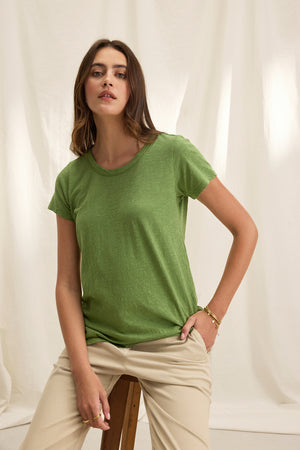 A woman in a Velvet by Graham & Spencer TILLY TEE and beige pants sits on a wooden stool against a light fabric background.