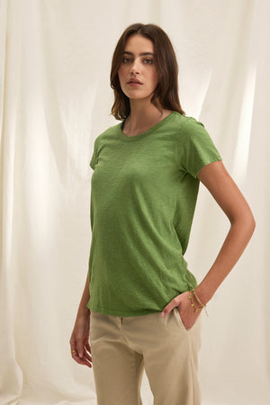 A woman stands against a beige backdrop wearing Velvet by Graham & Spencer's TILLY TEE in green and beige pants, embodying the easy-going charm of a California classic, with her hands by her sides.