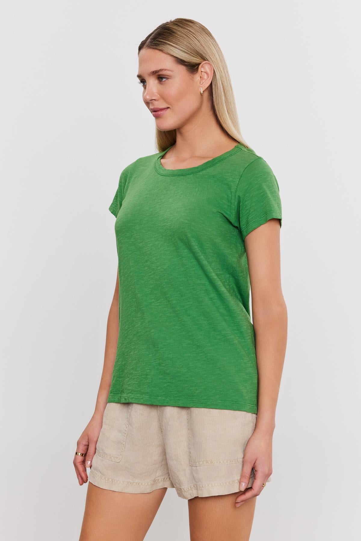   A woman with long blonde hair is wearing a green TILLY TEE by Velvet by Graham & Spencer and beige shorts, standing against a plain white background. She is looking to the side. 