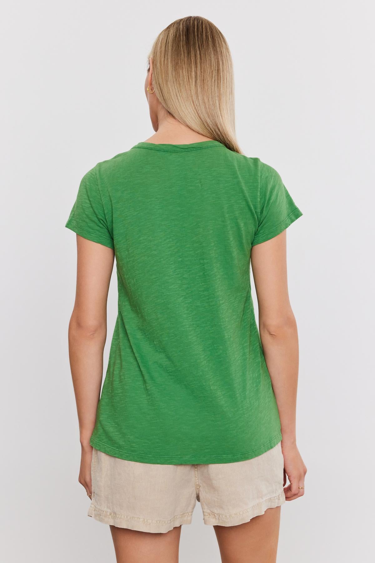   A person with long blonde hair is shown from behind, wearing the TILLY TEE in green by Velvet by Graham & Spencer and beige shorts. 