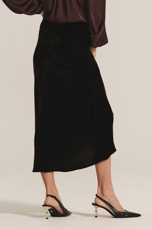 A person wears the FELICITY SKIRT by Velvet by Graham & Spencer—a dark brown silk velvet skirt with an elastic waistband—paired with a matching long-sleeve blouse and black slingback heels with gold accents, on a light background.