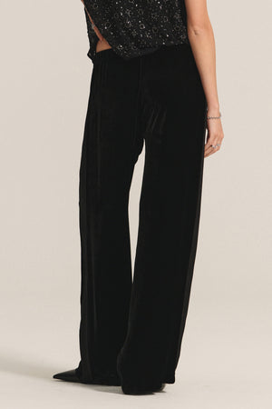 A person wearing the HILMA PANT by Velvet by Graham & Spencer—loose-fitting black silk velvet pants with a satin stripe—and a black sequined top, standing against a plain light background.