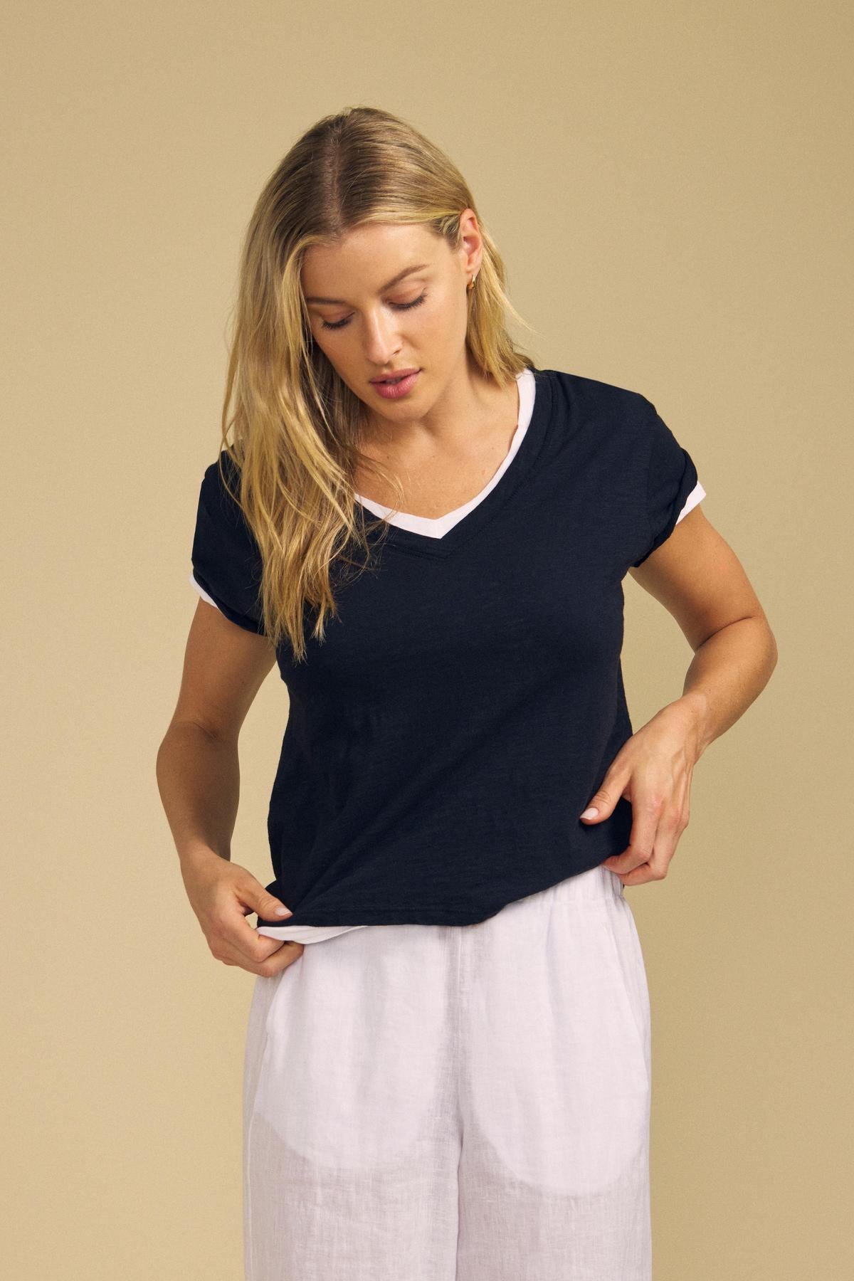 A woman wearing the ELISHA TEE by Velvet by Graham & Spencer—a navy V-neck tee with white trim—paired with white pants stands against a beige background, looking down and adjusting her shirt, showcasing this classic wardrobe staple perfect for effortless style.-42029084672193