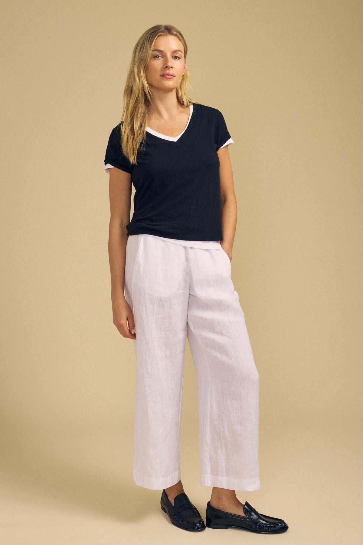 A woman stands against a beige background wearing the ELISHA TEE by Velvet by Graham & Spencer—a black cotton slub V-neck—paired with white wide-leg pants and black loafers. One hand rests in her pocket as she looks directly at the camera, showcasing an effortless look anchored by this timeless wardrobe staple.