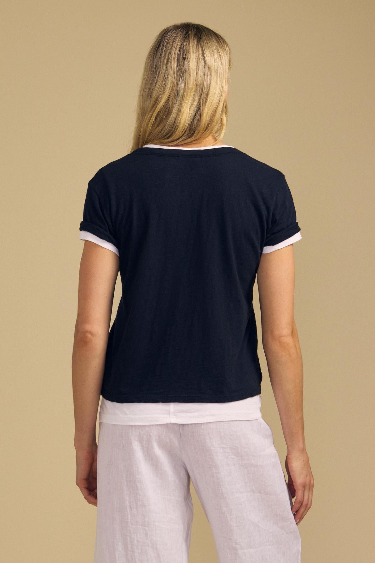 A person with long blonde hair is shown from behind wearing the ELISHA TEE by Velvet by Graham & Spencer—a black V-neck shirt with white trim and an effortless wardrobe staple—paired with white pants, standing against a plain beige background.-42029084639425