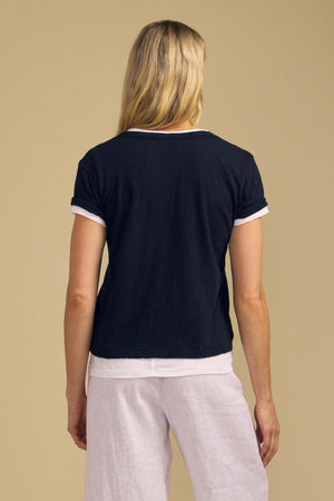 A person with long blonde hair is shown from behind wearing the ELISHA TEE by Velvet by Graham & Spencer—a black V-neck shirt with white trim and an effortless wardrobe staple—paired with white pants, standing against a plain beige background.