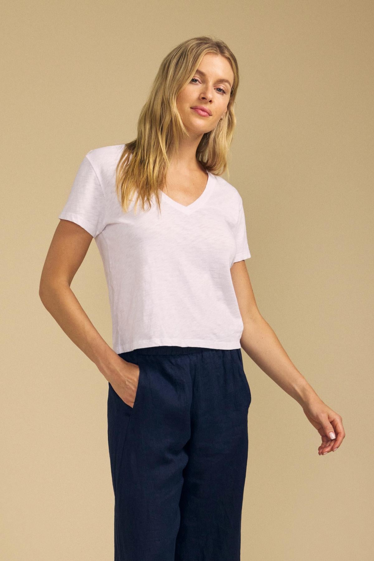 Dressed in the ELISHA TEE by Velvet by Graham & Spencer—a white cotton slub V-neck—paired with dark pants, a person stands against a beige background with one hand in their pocket, showcasing this timeless wardrobe staple.