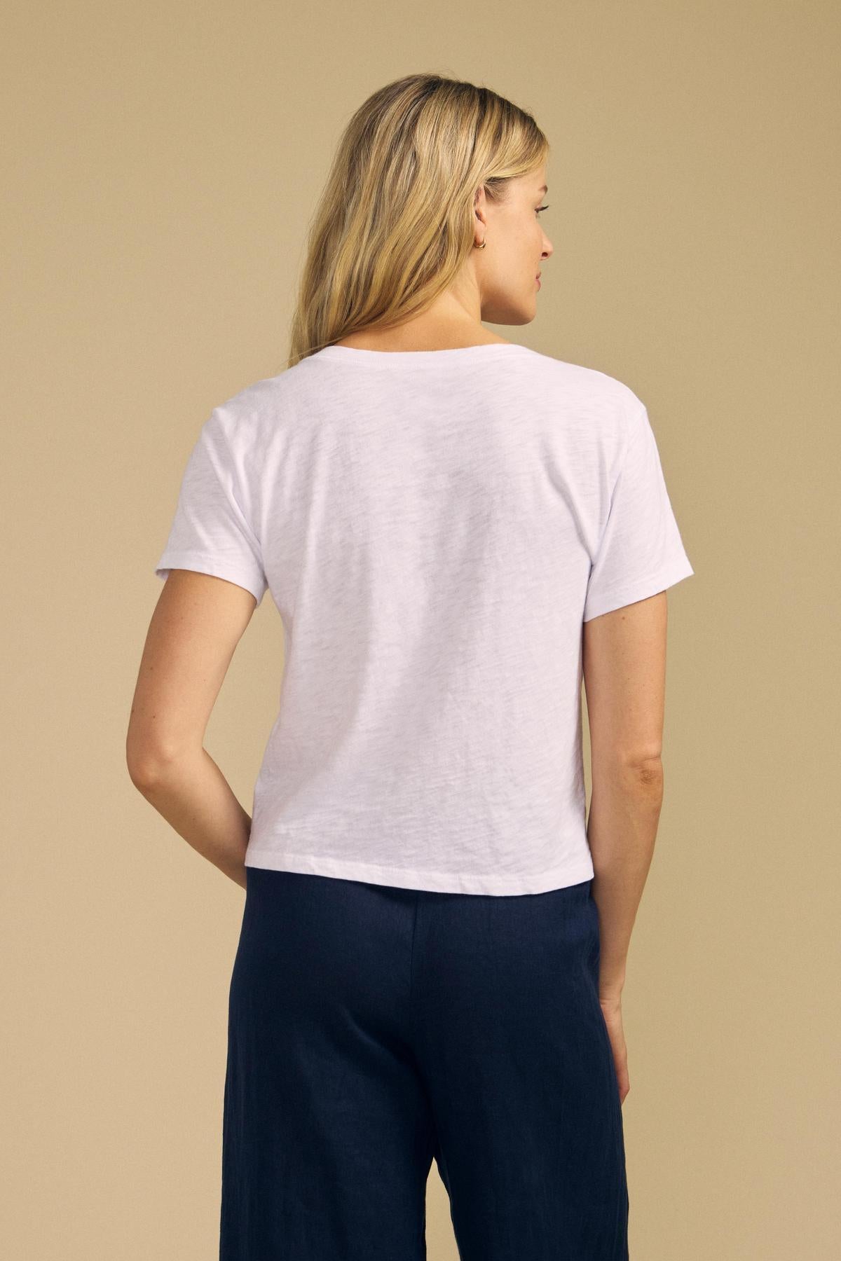 A person with long blonde hair, wearing the ELISHA TEE by Velvet by Graham & Spencer—a classic white cotton slub V-neck—paired with dark pants, stands facing away against a beige background.-42029091782849