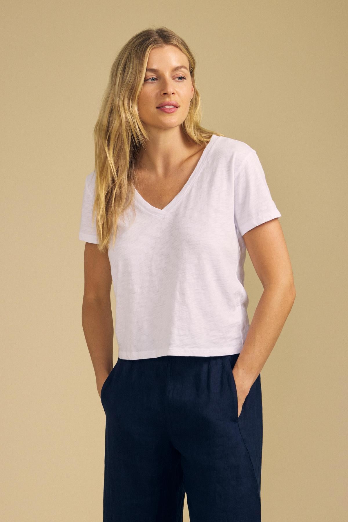 A woman stands with her hands in her pockets against a beige background, effortlessly showcasing the ELISHA TEE by Velvet by Graham & Spencer—a classic white V-neck, crafted from soft cotton slub—paired with navy pants for a timeless wardrobe staple.-42029091815617