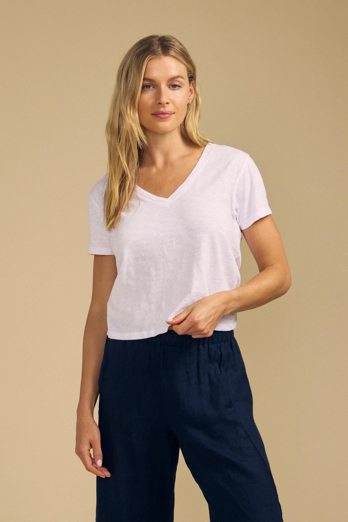 A woman with long blonde hair wears the classic ELISHA TEE by Velvet by Graham & Spencer—a white V-neck crafted from soft cotton slub—paired with dark blue pants, standing against a plain beige background.