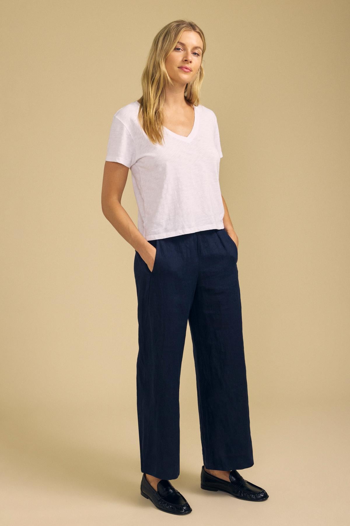 A person stands against a beige background wearing the ELISHA TEE by Velvet by Graham & Spencer—a classic white cotton slub V-neck—paired with dark wide-leg pants and black loafers, hands tucked casually in their pockets.-42029093552321