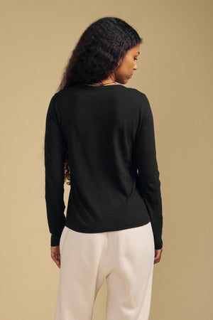 A person with long curly hair seen from behind is wearing the Velvet by Graham & Spencer LIZZIE TEE—a black, crew-neck, long-sleeve cotton slub top—paired with white pants and standing against a plain beige background.