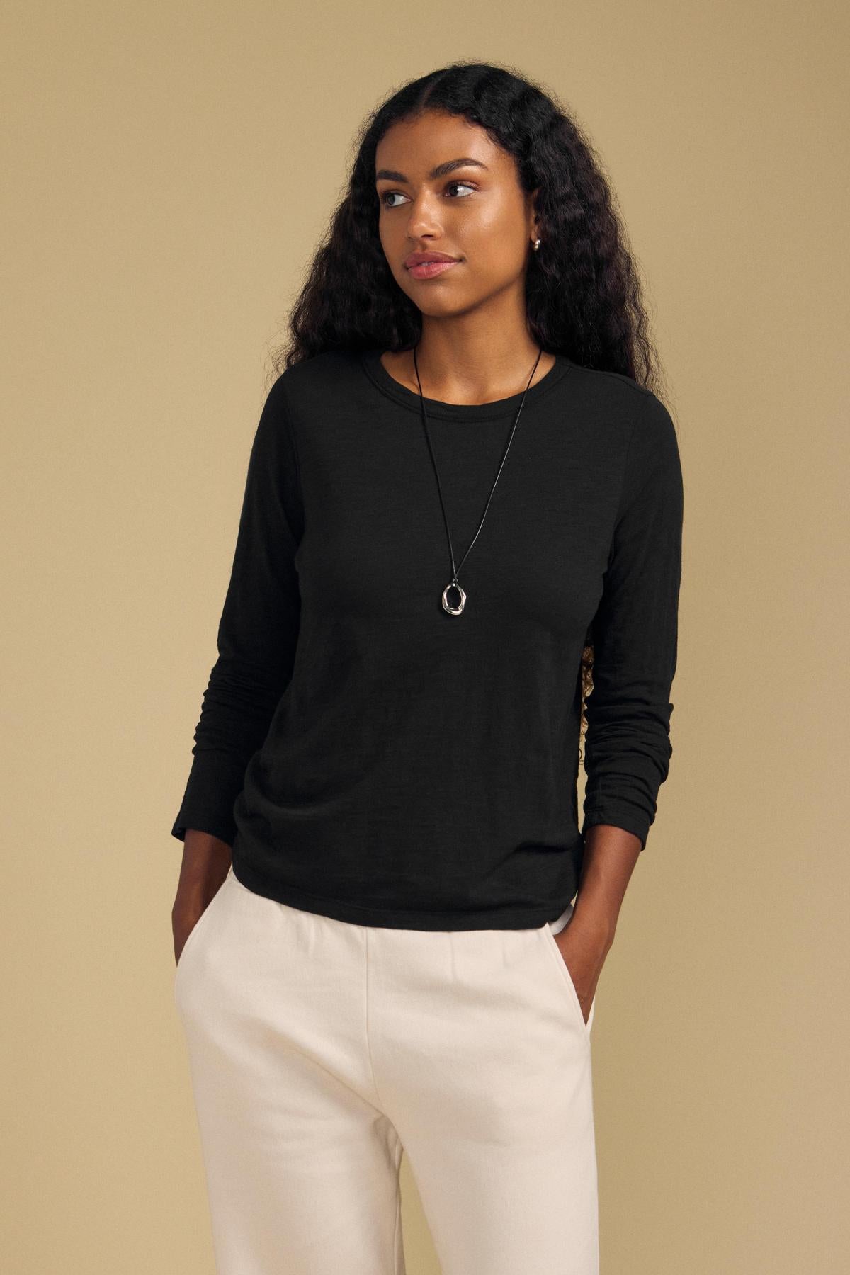 A woman with long curly hair stands against a beige background, wearing the Velvet by Graham & Spencer LIZZIE TEE—a black long sleeve top with a classic crew neckline—paired with white pants and a necklace, her hands in her pockets.