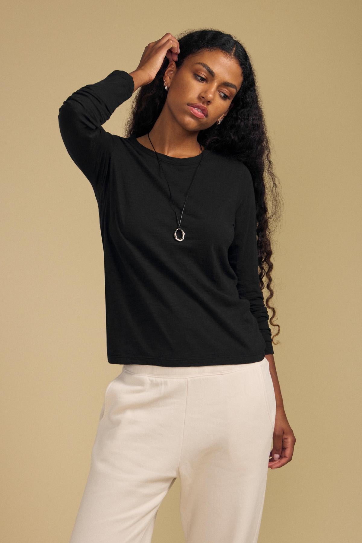 A woman wearing the LIZZIE TEE by Velvet by Graham & Spencer—a black long-sleeve cotton slub top with a classic crew neckline—paired with cream pants, stands against a beige background, one hand touching her hair and the other at her side.-42029100892353