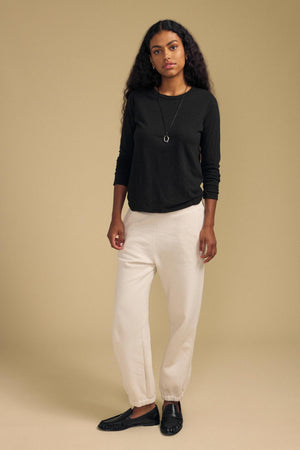 A woman stands against a beige background, wearing the LIZZIE TEE by Velvet by Graham & Spencer—a black long sleeve cotton slub top with a crew neckline—paired with white relaxed pants, black shoes, and a necklace.