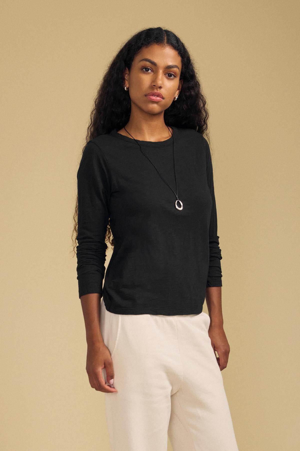 A woman with long curly hair wears the Velvet by Graham & Spencer LIZZIE TEE, a black long sleeve cotton slub top with a crew neckline, paired with light pants and a pendant necklace, standing against a beige background.