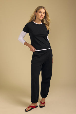 A woman stands against a beige background, wearing the MARIKA TEE by Velvet by Graham & Spencer—a boxy fit black tee crafted from soft cotton slub—layered over a white long-sleeve top, paired with black jogger pants and red sandals.