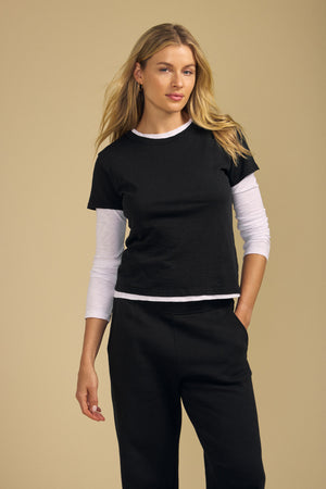 A woman stands against a plain beige background, wearing the MARIKA TEE—a black short-sleeve T-shirt by Velvet by Graham & Spencer, crafted from soft cotton slub—layered over a white long-sleeve shirt and paired with black pants, with one hand in her pocket. The boxy fit of the tee adds a relaxed yet modern touch to her look.