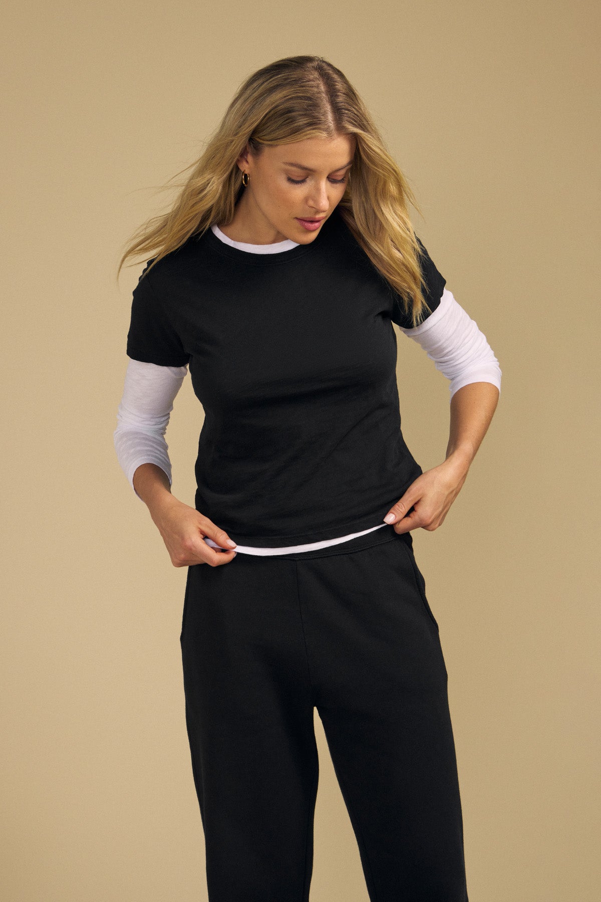 A woman with long blonde hair wears the MARIKA TEE by Velvet by Graham & Spencer—a black cotton slub T-shirt in a relaxed boxy fit—layered over a white long-sleeve shirt and paired with black pants, standing against a beige background.