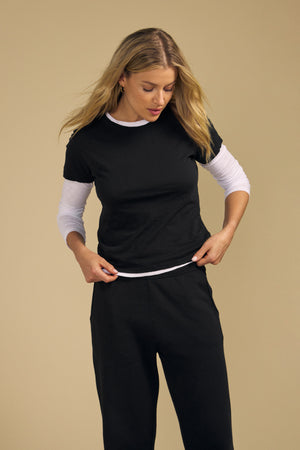 A woman with long blonde hair wears the MARIKA TEE by Velvet by Graham & Spencer—a black cotton slub T-shirt in a relaxed boxy fit—layered over a white long-sleeve shirt and paired with black pants, standing against a beige background.