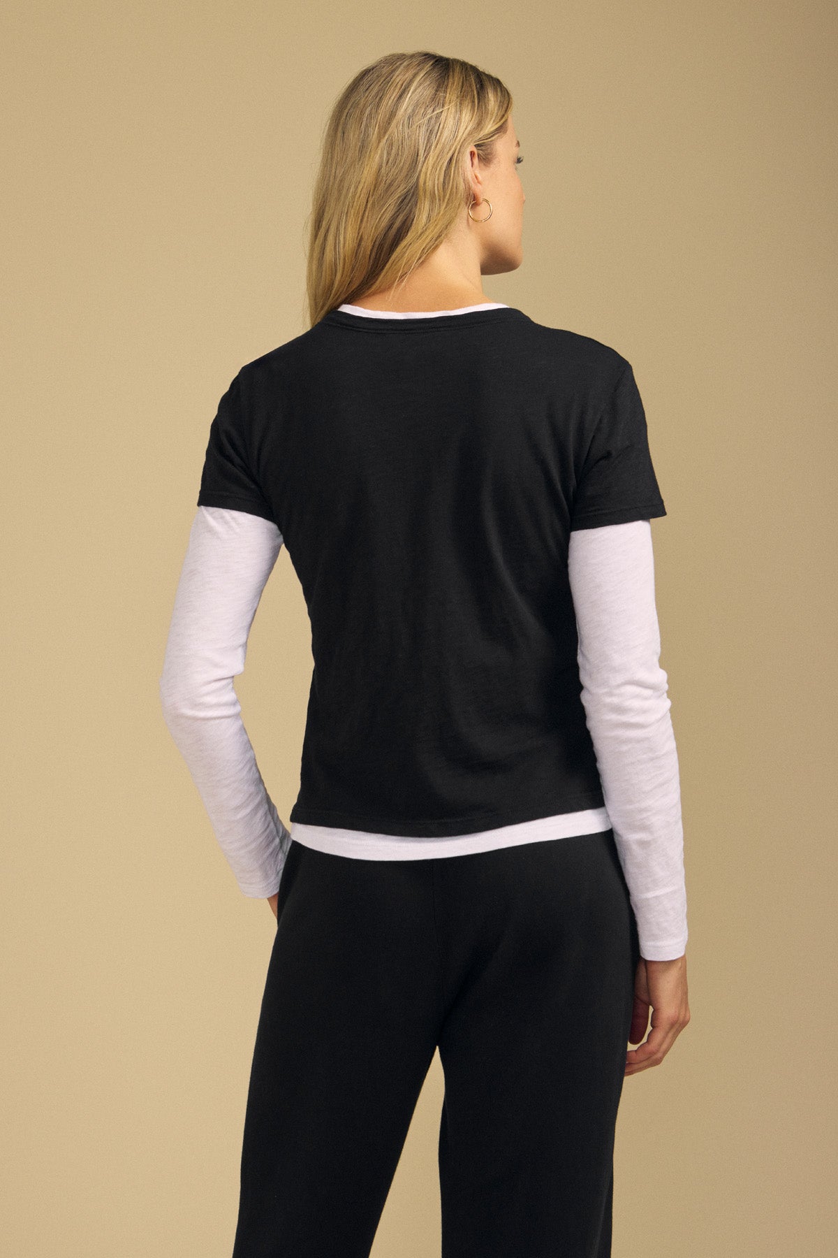 A person stands with their back to the camera, wearing the boxy fit black MARIKA TEE by Velvet by Graham & Spencer layered over a long-sleeve white shirt and paired with black pants, set against a beige background.
