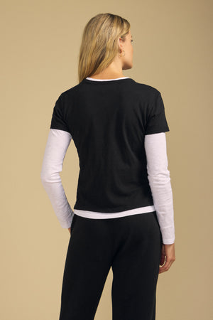 A person stands with their back to the camera, wearing the boxy fit black MARIKA TEE by Velvet by Graham & Spencer layered over a long-sleeve white shirt and paired with black pants, set against a beige background.