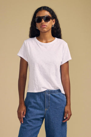 A woman wearing sunglasses and blue jeans stands against a beige background with her left hand in her pocket, dressed in the MARIKA TEE by Velvet by Graham & Spencer—a plain white cotton slub tee featuring a relaxed boxy fit.