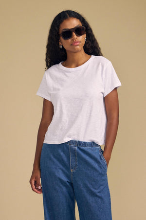 A woman wearing sunglasses, blue jeans, and the MARIKA TEE—a relaxed, boxy-fit white cotton slub top by Velvet by Graham & Spencer—stands against a beige background with her left hand in her pocket.