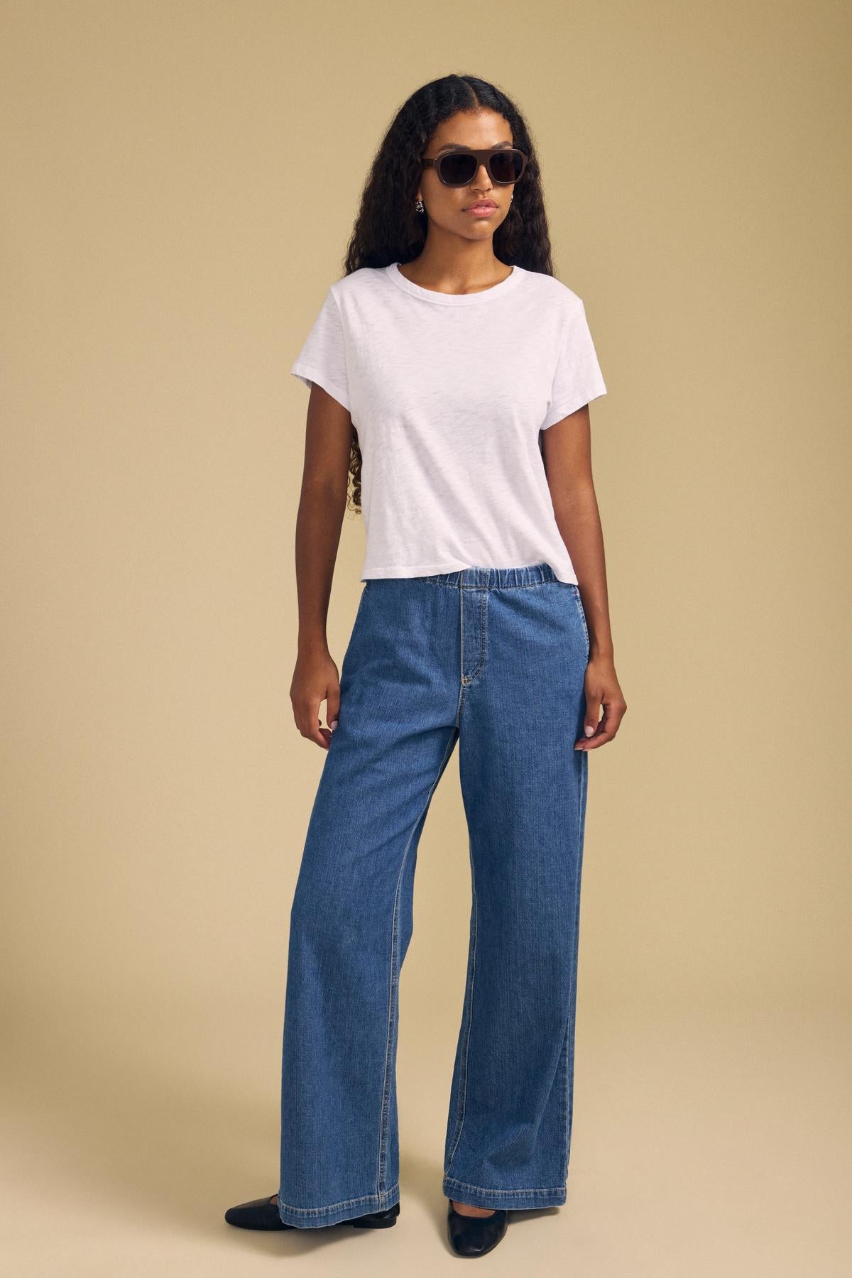 A woman wearing sunglasses, the MARIKA TEE by Velvet by Graham & Spencer—a plain white cotton slub tee with a relaxed boxy fit—paired with wide-leg blue jeans and black shoes, stands against a beige background.