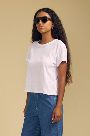 A woman wearing sunglasses and blue jeans stands against a beige background, facing slightly to the side with her arms relaxed at her sides. She wears the MARIKA TEE by Velvet by Graham & Spencer—a plain white, soft cotton slub top with a relaxed boxy fit that gives her look an effortless vibe.