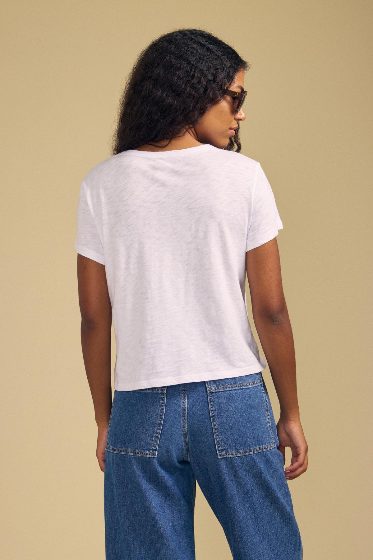 A person with long curly hair wearing sunglasses and a relaxed, boxy fit MARIKA TEE in cotton slub by Velvet by Graham & Spencer, paired with blue jeans, stands with their back to the camera against a beige background.