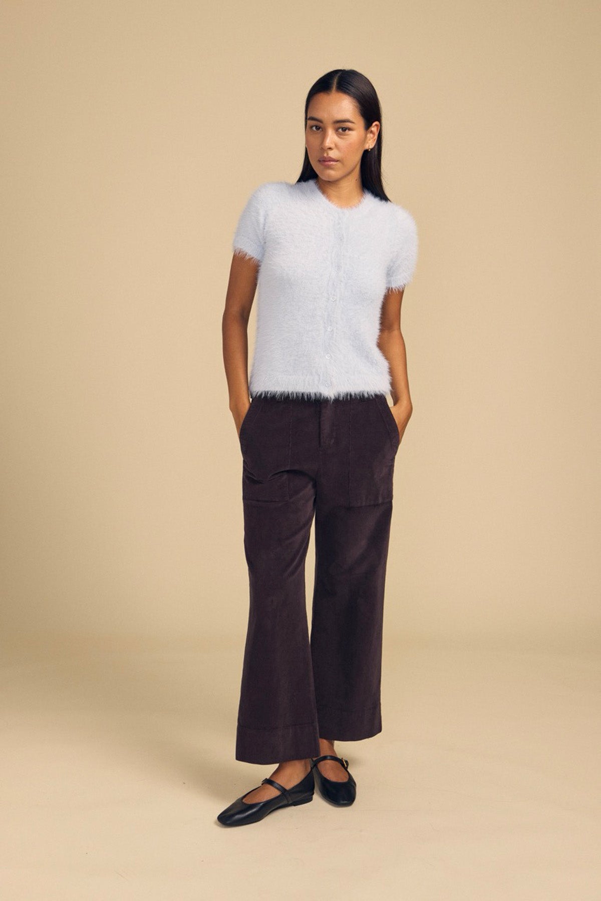 A woman wears a light blue fuzzy short-sleeved sweater and dark brown VERA CORDUROY PANT, which are high-rise wide-leg trousers featuring front patch pockets.