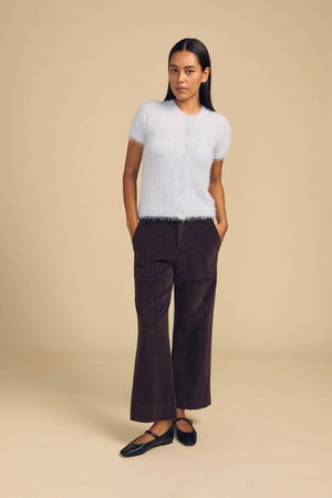 A woman wears a light blue fuzzy short-sleeved sweater and dark brown VERA CORDUROY PANT, which are high-rise wide-leg trousers featuring front patch pockets.