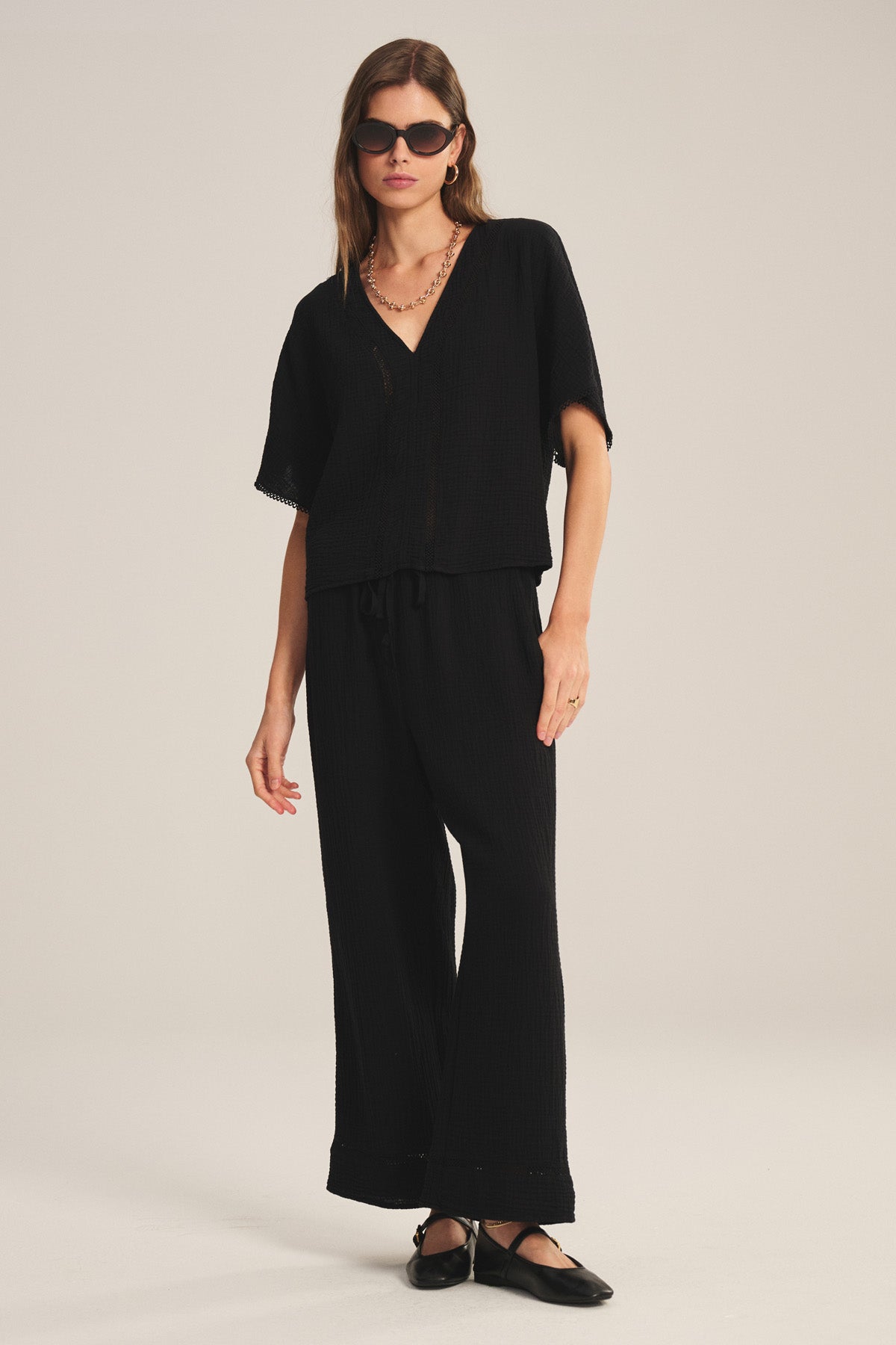 A woman stands in a studio wearing the VERNA TOP by Velvet by Graham & Spencer—a loose-fitting black deep V-neck cotton gauze blouse—paired with matching wide-leg pants, black ballet flats, sunglasses, a chain necklace, and a ring.
