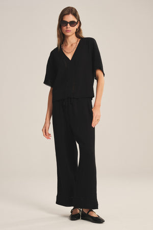 A woman stands in a studio wearing the VERNA TOP by Velvet by Graham & Spencer—a loose-fitting black deep V-neck cotton gauze blouse—paired with matching wide-leg pants, black ballet flats, sunglasses, a chain necklace, and a ring.