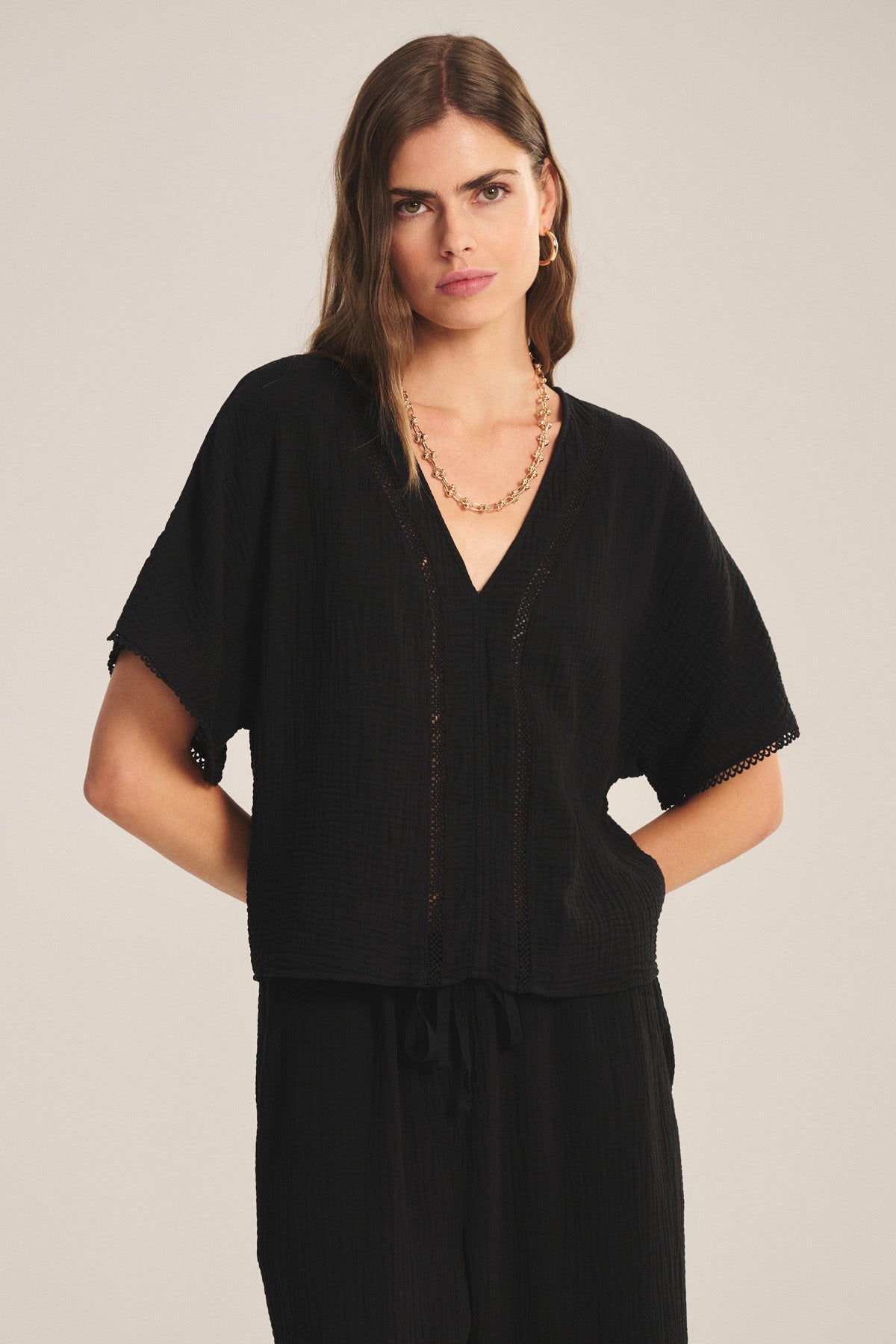 A woman models the Velvet by Graham & Spencer VERNA TOP—a black short-sleeve deep V-neck with subtle lace cutouts—paired with black pants, standing against a plain background and looking at the camera, hands on her hips.