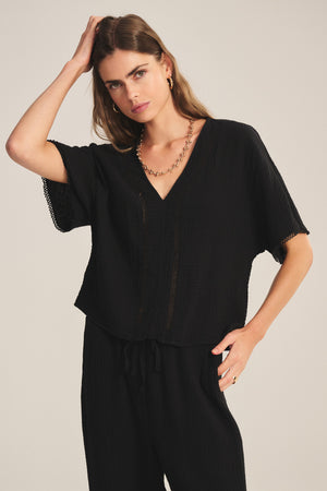 A woman wears the VERNA TOP by Velvet by Graham & Spencer, a loose-fitting black cotton gauze top with a deep V-neck, paired with matching black pants while posing facing the camera with one hand on her head and the other on her hip.
