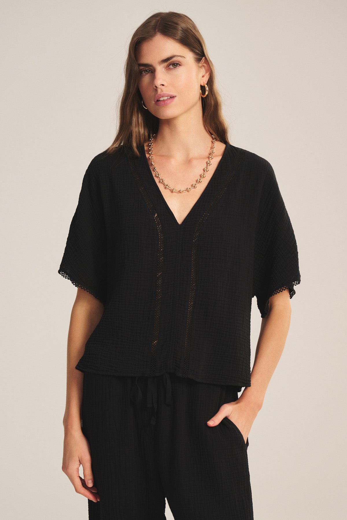 A woman wears the VERNA TOP by Velvet by Graham & Spencer, a loose black cotton gauze piece with a deep V-neck and short sleeves, paired with black pants and a gold chain necklace.