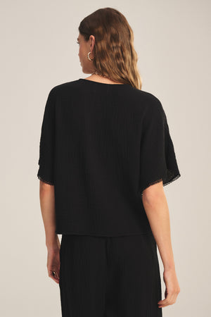 A woman with wavy brown hair wears the Velvet by Graham & Spencer VERNA TOP, a loose black cotton gauze blouse, paired with matching pants. She is shown from the back against a neutral background.
