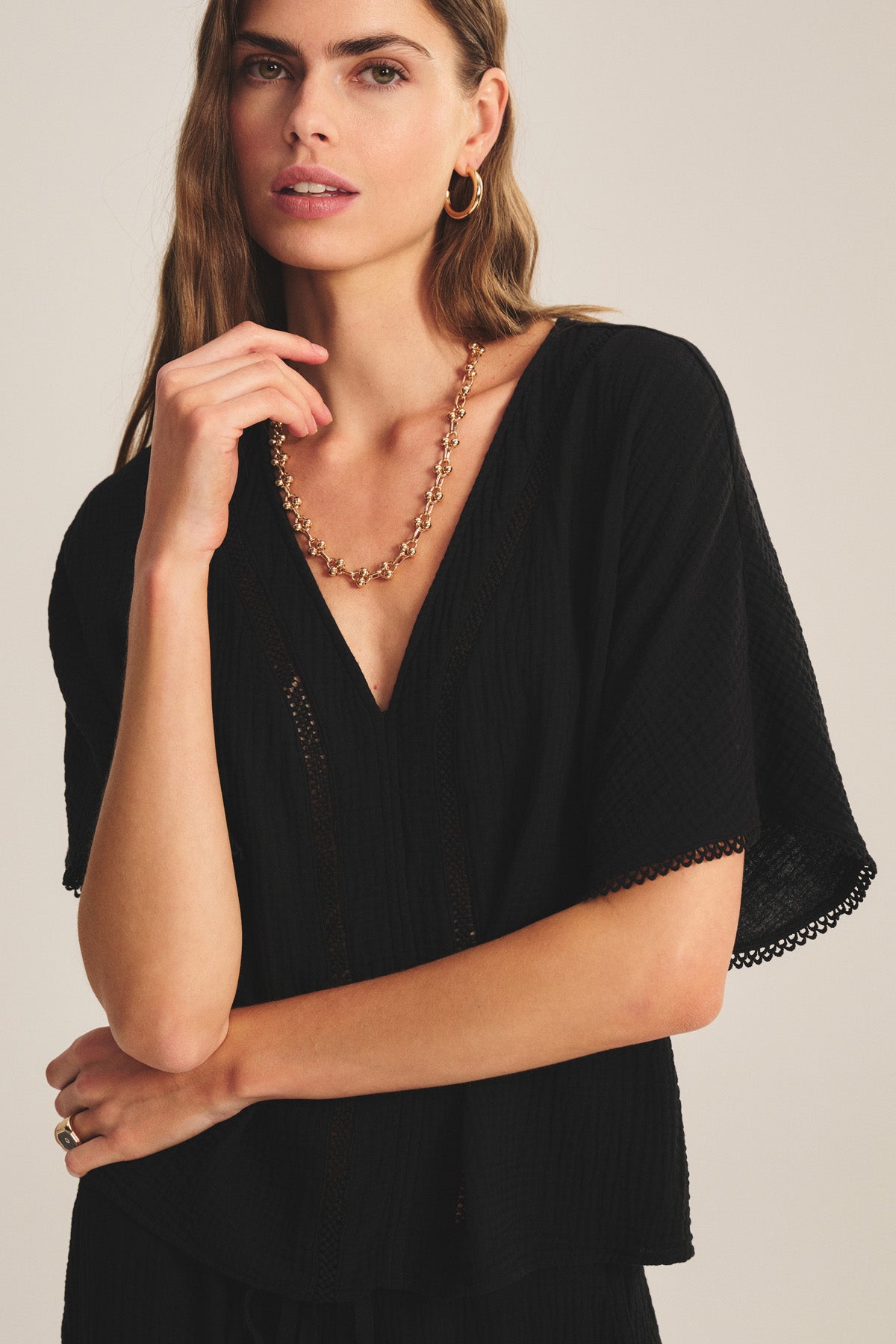 Woman with long hair wearing the VERNA TOP by Velvet by Graham & Spencer—a black cotton gauze top with a deep V-neck—paired with gold hoop earrings and a gold chain necklace, standing against a neutral background.-41297002725569