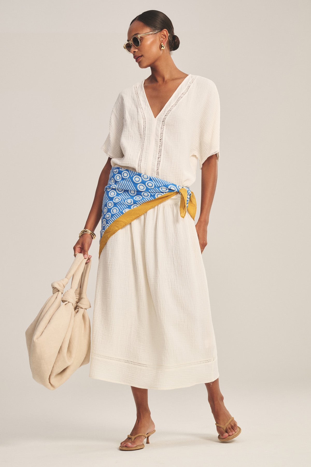 Woman wearing the VERNA TOP by Velvet by Graham & Spencer, paired with a blue and yellow scarf belt, tan heeled sandals, sunglasses, and carrying a large beige bag.